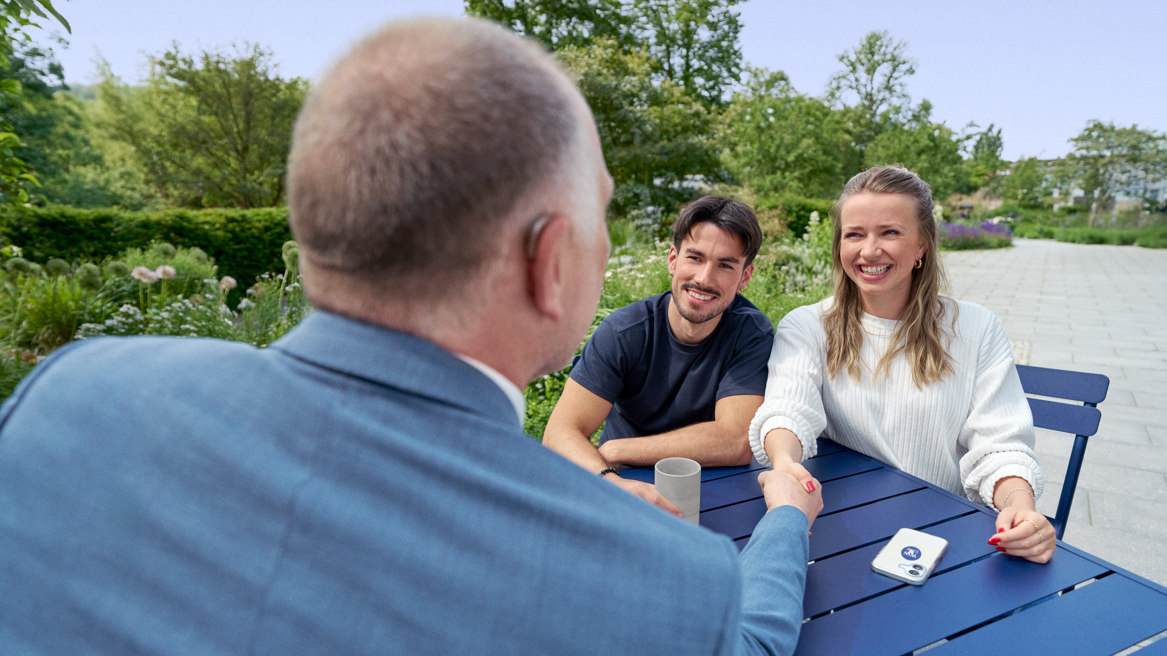 Three colleagues of different generations talking at a table, two shaking hands, all smiling.