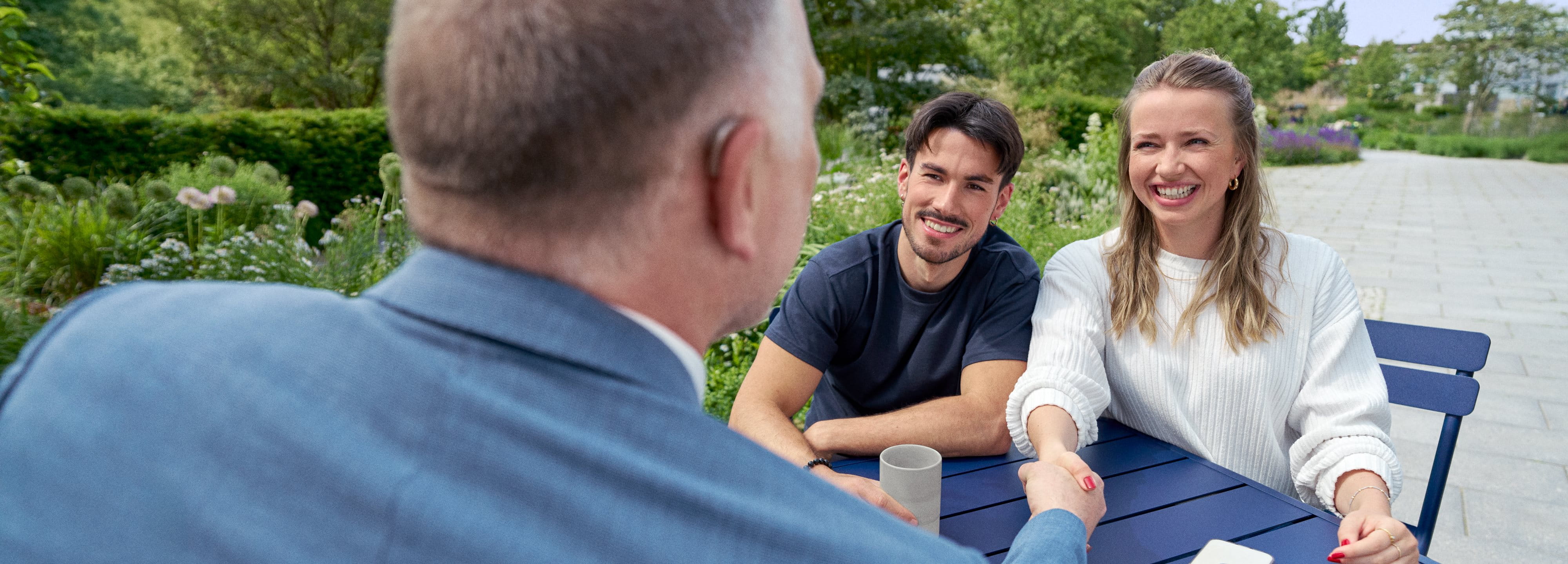 Three colleagues of different generations talking at a table, two shaking hands, all smiling.