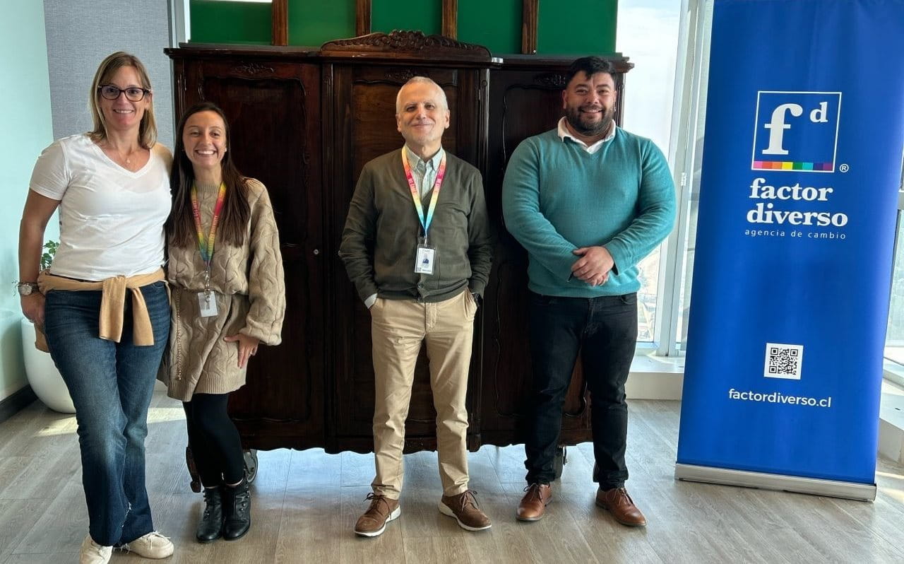 Vier Personen stehen in einem modernen Büro mit großen Fenstern und Hängelampen vor einem großen Holzschrank, daneben ein blauer Banner mit dem Logo und der Aufschrift „factor diverso“ auf Spanisch.