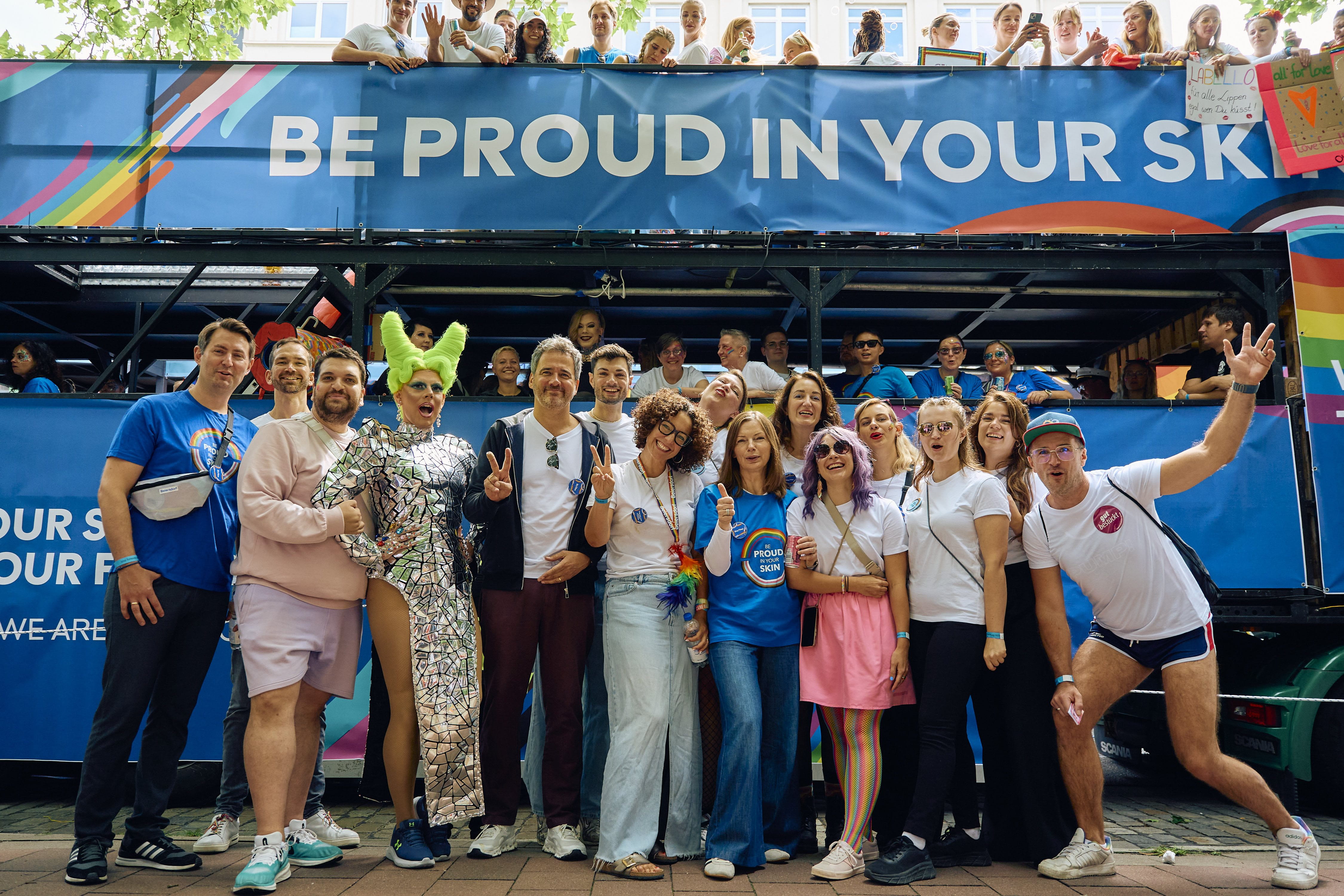 Eine Gruppe von Personen posiert und jubelt vor einem Pride-Truck, der mit Regenbogenfarben und dem Slogan „BE PROUD IN YOUR SKIN“ dekoriert ist; auf der oberen Ebene des Trucks sind weitere Teilnehmende zu sehen.