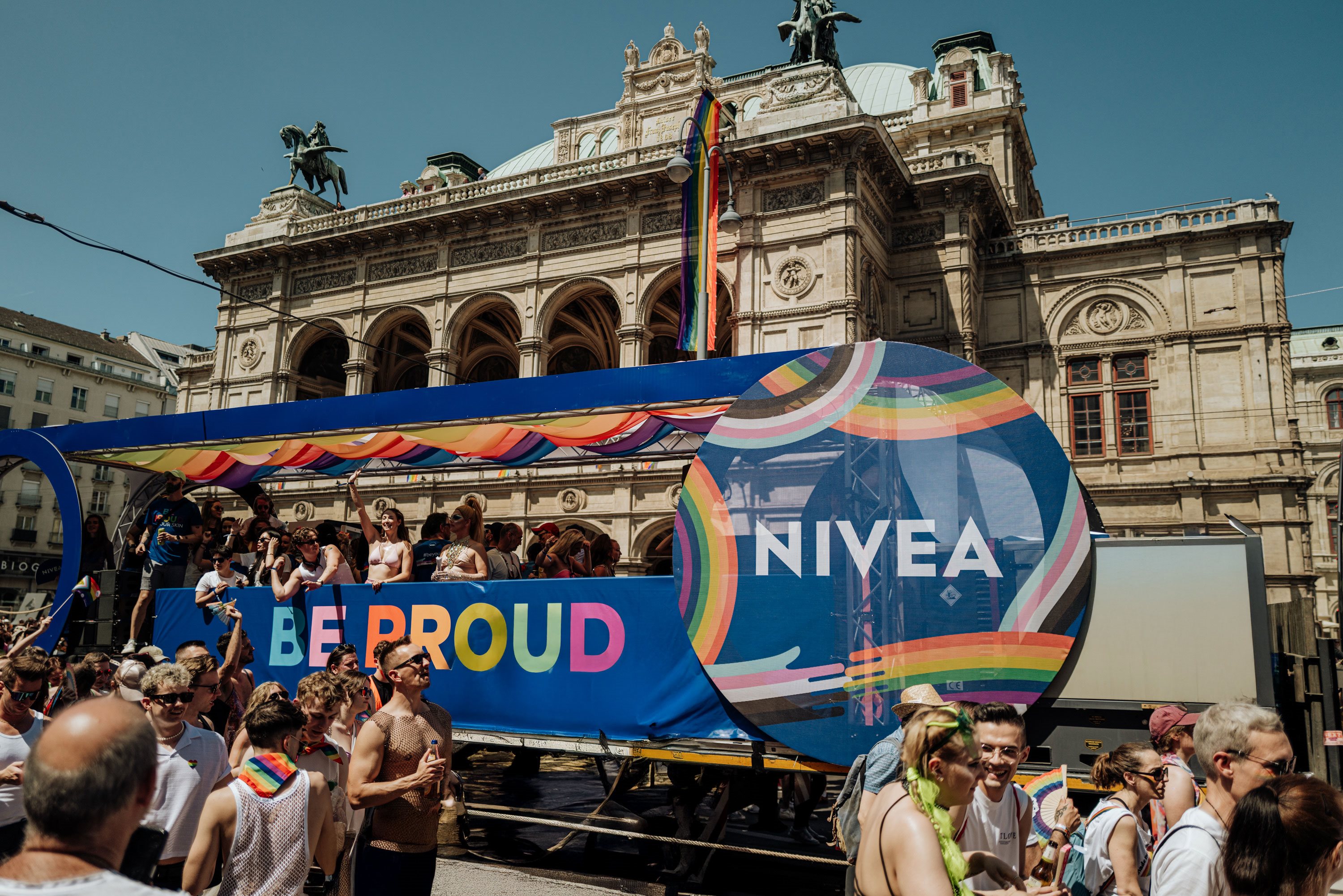 Ein bunter, NIVEA-gebrandeter Pride-Truck mit dem Slogan „BE PROUD“ in Regenbogenfarben, umgeben von feiernden Menschen vor einem historischen Gebäude mit Regenbogenflaggen.