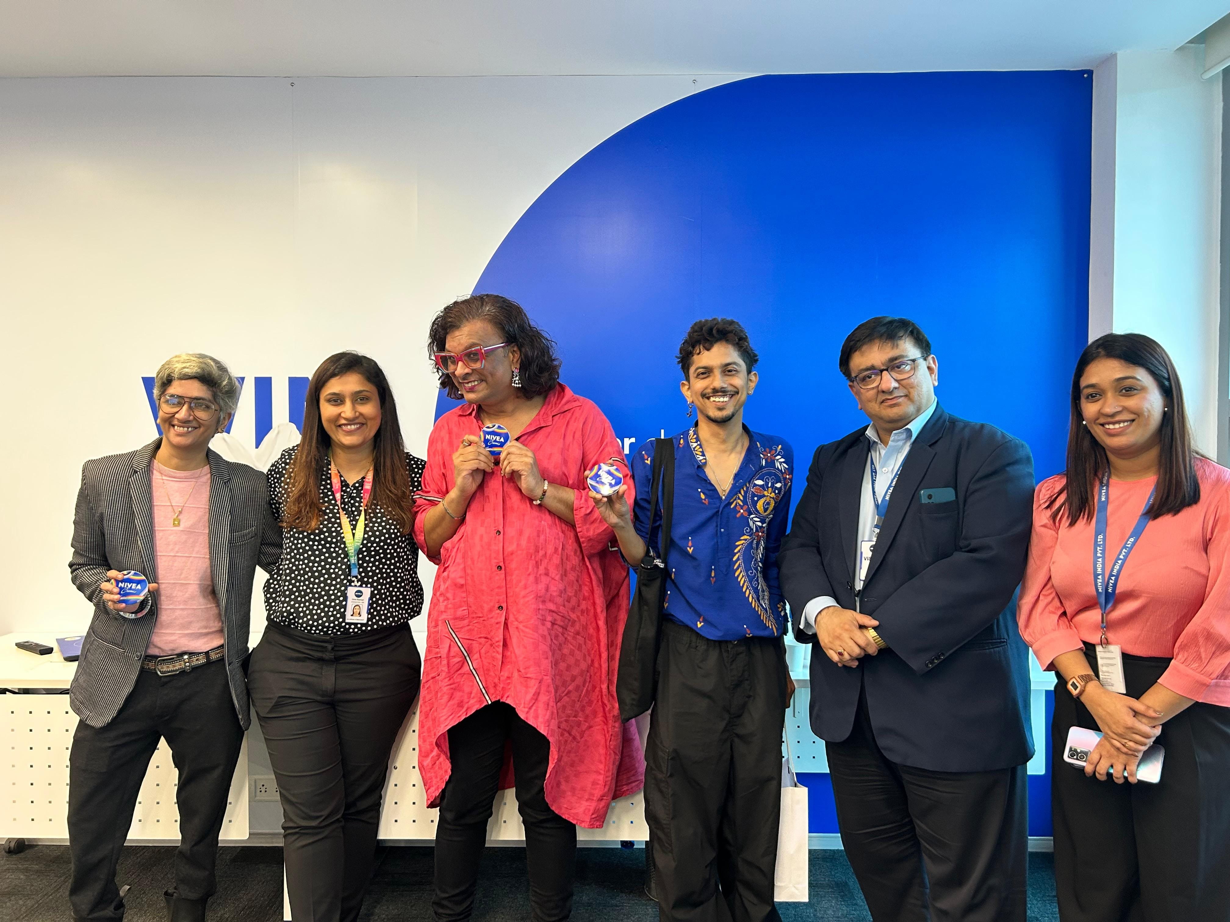 Personen in blauen T-Shirts mit der Aufschrift “Proud in Your Skin” stehen vor einem weißen Zelt bei einem Event im Park, im Hintergrund sind weitere Besucher*innen zu sehen.