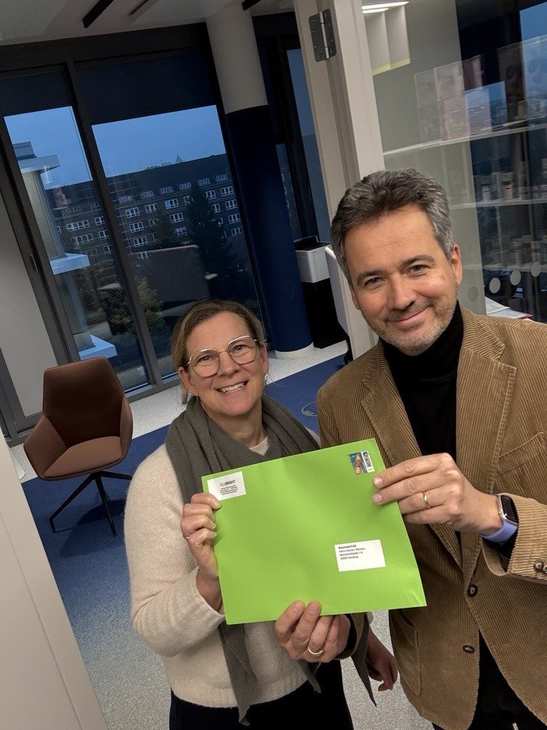 Zwei Personen stehen in einem Innenraum, lächeln und halten gemeinsam einen leuchtend grünen Umschlag hoch; im Hintergrund sind Bürofenster und Möbel zu sehen.