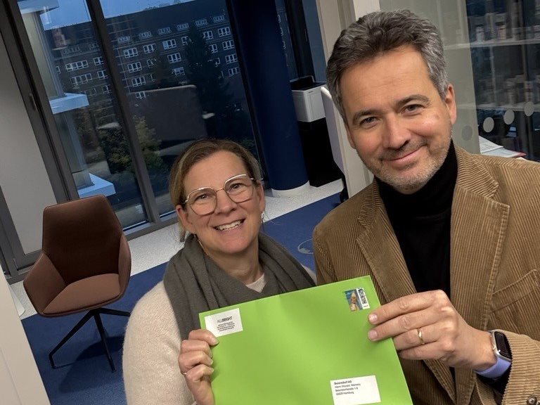 Zwei Personen stehen in einem Innenraum, lächeln und halten gemeinsam einen leuchtend grünen Umschlag hoch; im Hintergrund sind Bürofenster und Möbel zu sehen.