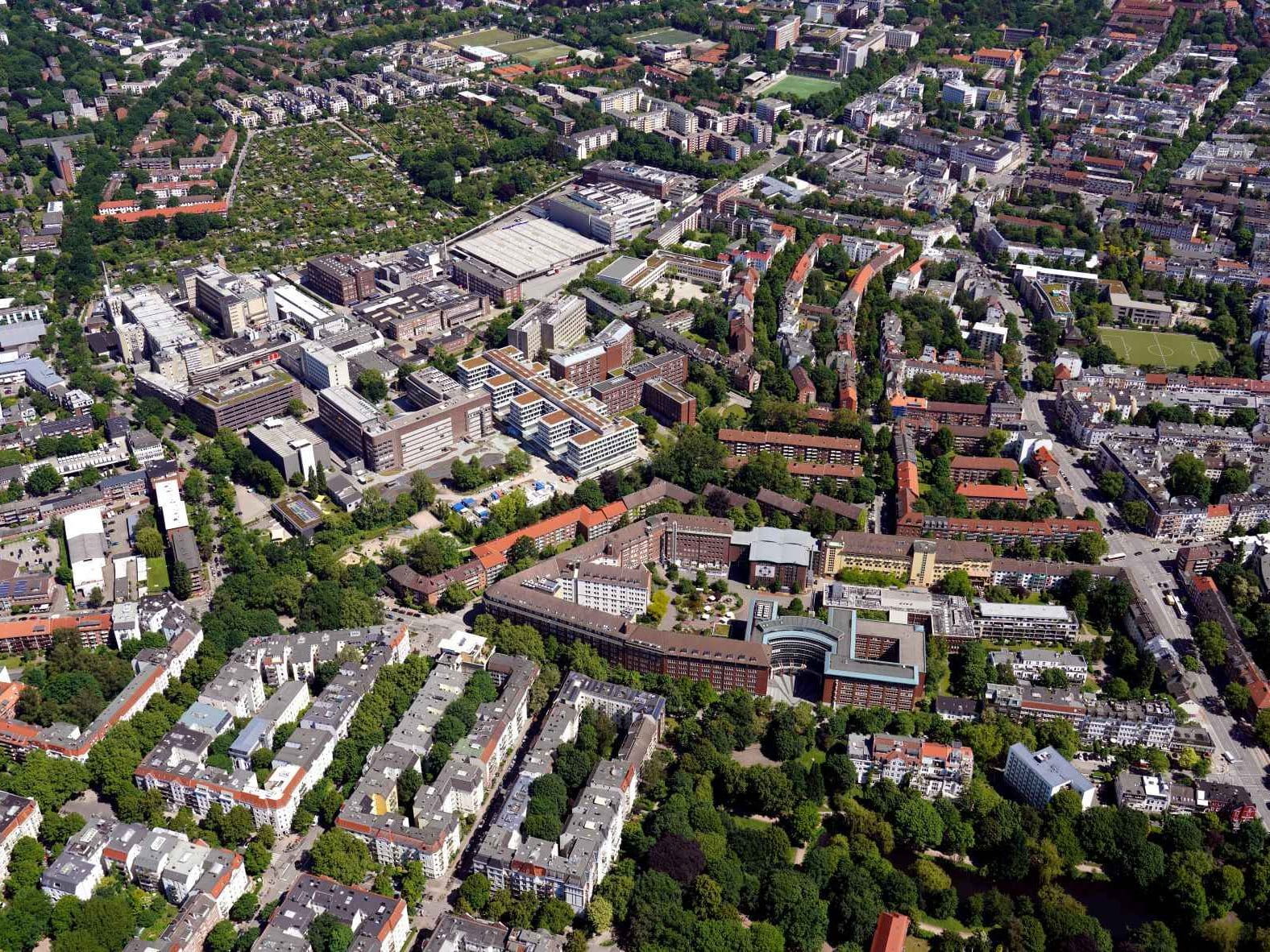 An aerial photograph showing a dense urban landscape of a city, with a large industrial or corporate complex composed of various brick and modern buildings at its center. The surrounding area is a mix of residential apartment blocks with red-tiled roofs and numerous green spaces filled with mature trees. Roads and streets wind through the neighborhood, with several larger buildings and courtyards visible throughout the expansive city view under clear daylight.