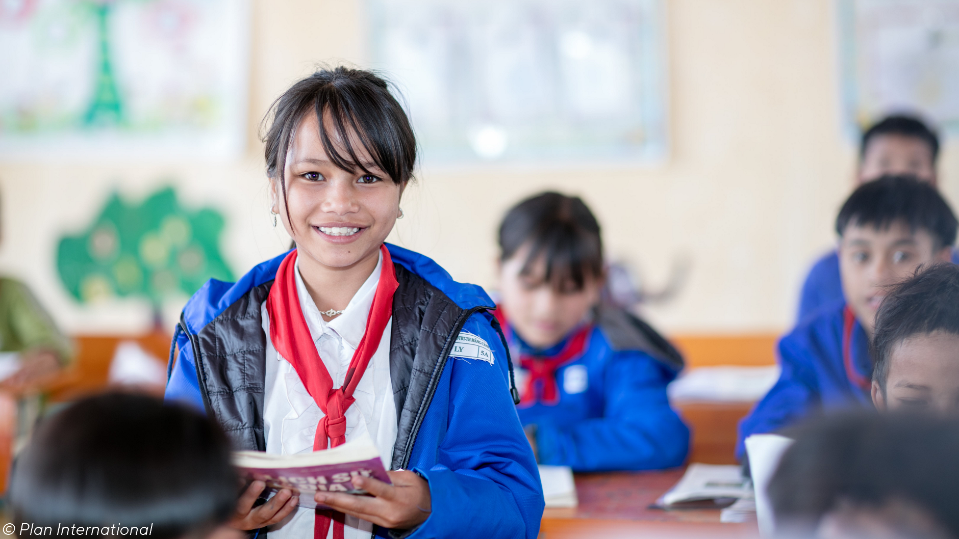Das Bild zeigt eine latein-amerikanische Schulklasse, in der die Schüler und Schülerinnen alle dieselbe Uniform tragen. Im Vordergrund ist eine junge, lächelnde Schülerin zu sehen, die eine blaue Jacke und einen roten Schal trägt und ein Buch oder ein Notizbuch in der Hand hält. Andere Schüler ihren Alters sitzen an ihren Plätzen um sie herum. Die Klassenzimmerumgebung wirkt hell und farbenfroh mit Kunstwerken an den Wänden. 