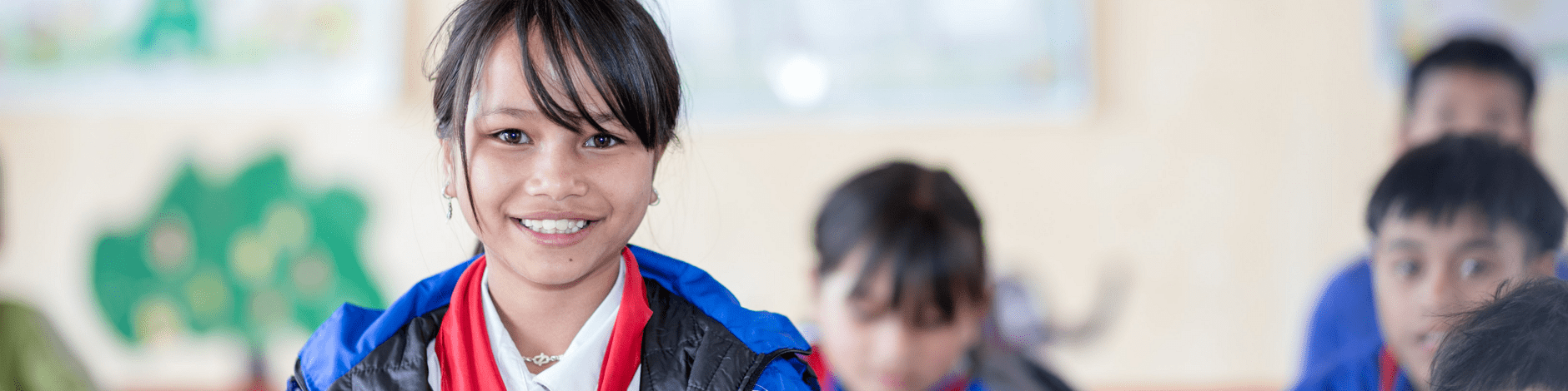 Das Bild zeigt eine latein-amerikanische Schulklasse, in der die Schüler und Schülerinnen alle dieselbe Uniform tragen. Im Vordergrund ist eine junge, lächelnde Schülerin zu sehen, die eine blaue Jacke und einen roten Schal trägt und ein Buch oder ein Notizbuch in der Hand hält. Andere Schüler ihren Alters sitzen an ihren Plätzen um sie herum. Die Klassenzimmerumgebung wirkt hell und farbenfroh mit Kunstwerken an den Wänden.