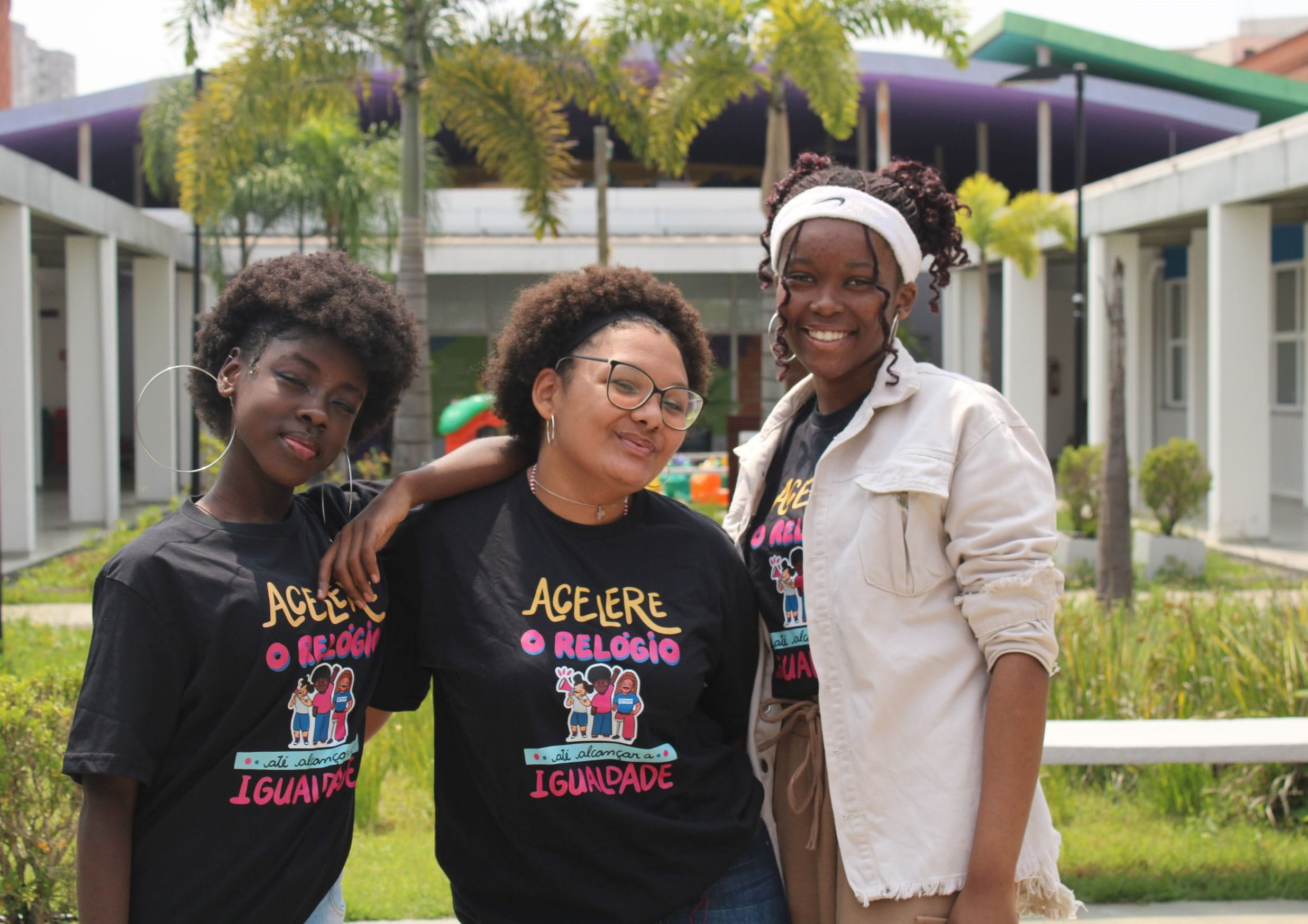3 junge Frauen aus Brasilien mit dunklem Hautton posieren lächelnd und Arm in Arm eng nebeneinanderstehend vor einem Gebäude mit Grünanlage. Sie tragen alle das gleiche, dunkle T-Shirt mit Bunter Aufschrift. 