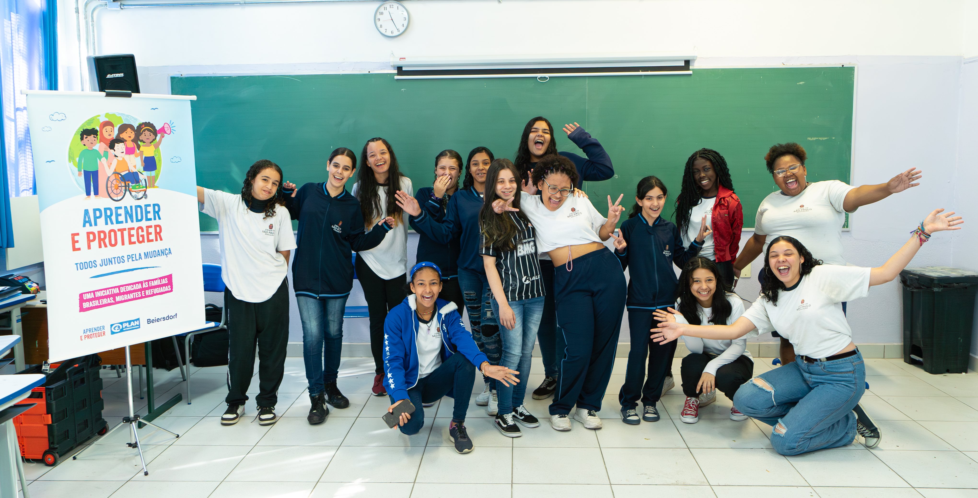 Ein Gruppen Foto von Mädchen aus Brasilien die fröhlich in einem Klassenzimmer vor einer grünen Tafel posieren. Die Gruppe posiert neben einem Plakat auf einem Aufsteller, auf dem Text und Cartoon Bilder, die Kinder abbilden, zu sehen ist. 