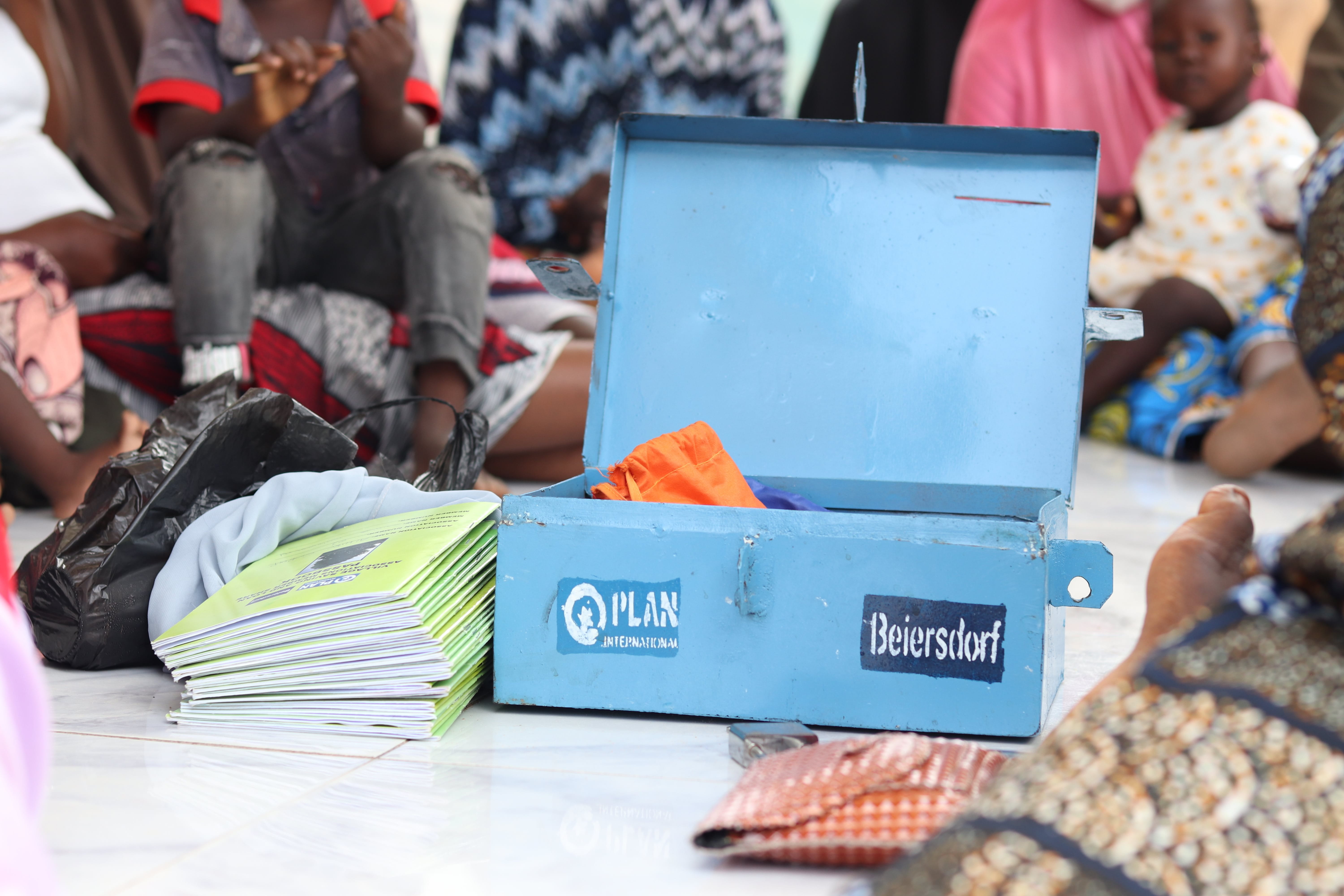 Eine blaue, offene Box mit den Logos von Plan International und Beiersdorf auf der Vorderseite. Die Box liegt in der Mitte eines Kreises aus Menschen mit dunklen Hautton, die um die Box herumsitzen, die aber nur zu erahnen sind, da der Fokus auf der Box liegt und der Rest verschwommen ist. 