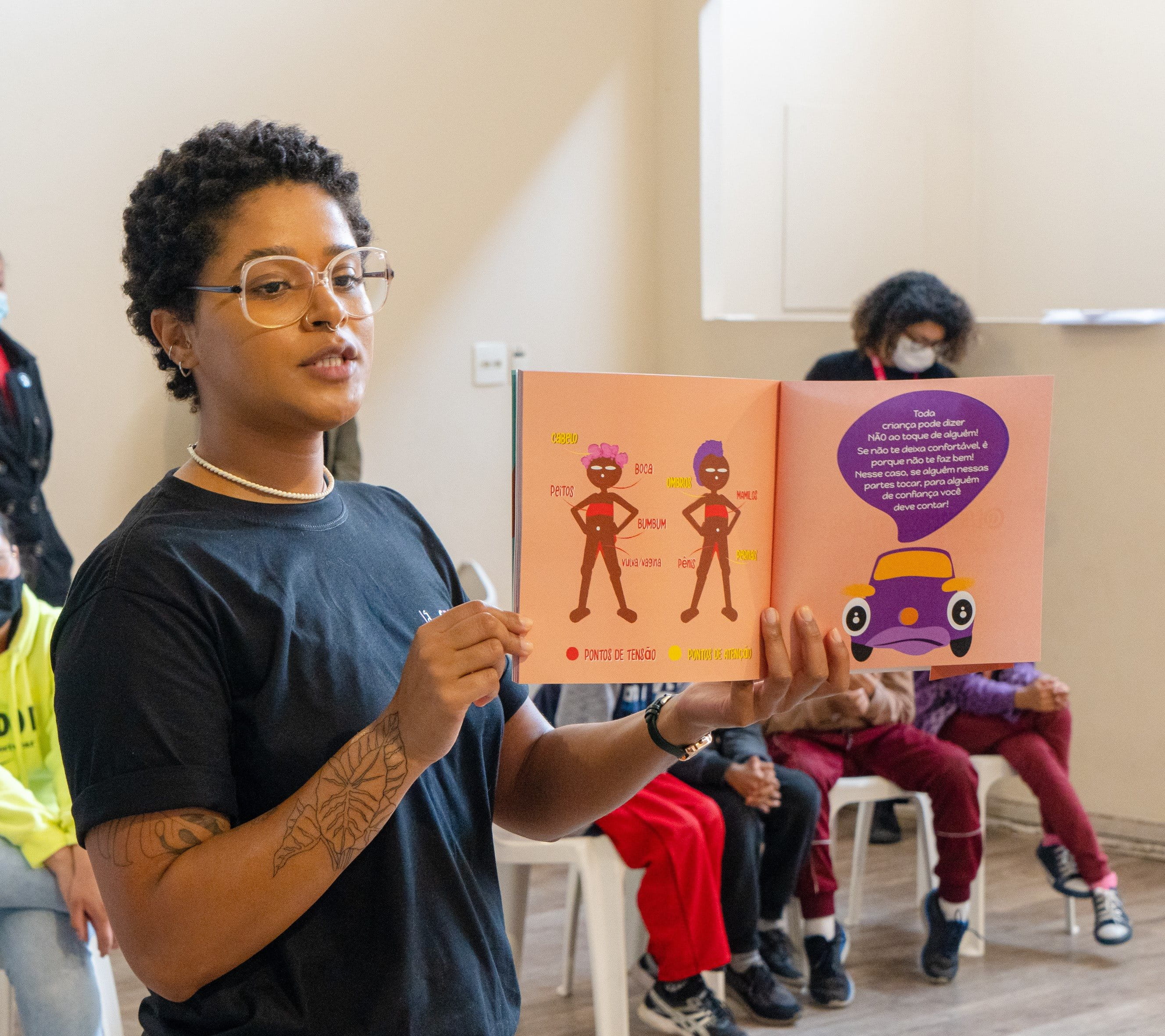 Das Bild zeigt eine Szene, in der eine Person ein aufgeschlagenes Kinderbuch mit farbenfrohen Illustrationen und Text hält. Die Zuhörer sitzen in einem hellen Raum und betrachten aufmerksam das Buch. Die Illustrationen im Buch zeigen zwei Charaktere und ein Auto. Die Person, die das Buch hält, trägt ein dunkles T-Shirt und hat Tätowierungen auf den Armen. Die Umgebung ist freundlich und gut beleuchtet, mit weißen Wänden und Holzboden.