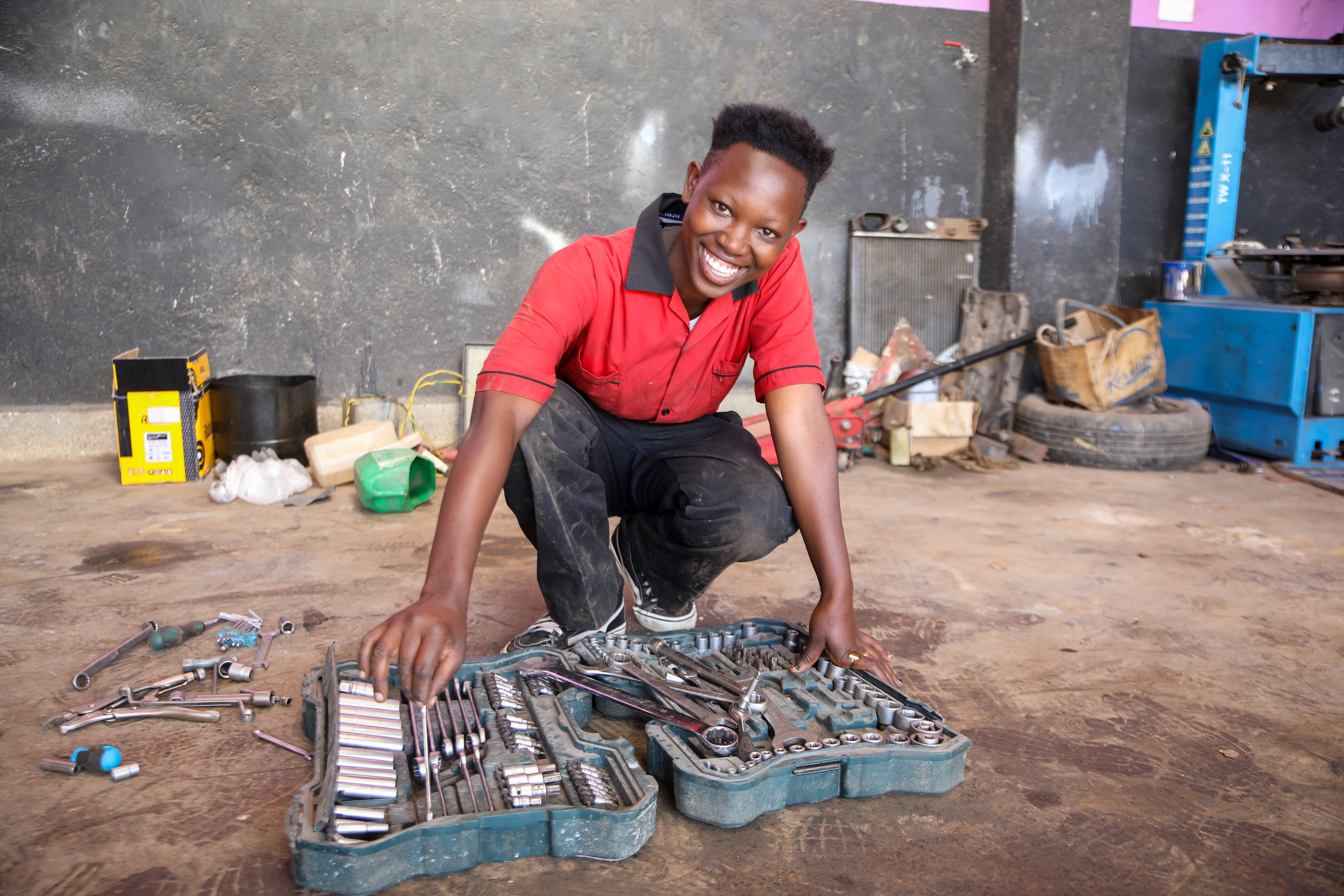 Eine Mechanikerin mit dunklem Hautton, die auf dem Boden kniet und Werkzeuge in einer offenen Werkzeugkiste sortiert. Die Werkzeuge umfassen verschiedene Arten von Schraubenschlüsseln und anderen Handwerkzeugen. Im Hintergrund sind eine dunkle Wand sowie verschiedene Gegenstände wie Ölkanister und ein Reifen zu sehen. Der Boden besteht aus Beton und weist verschiedene Flecken und Verschmutzungen auf. 