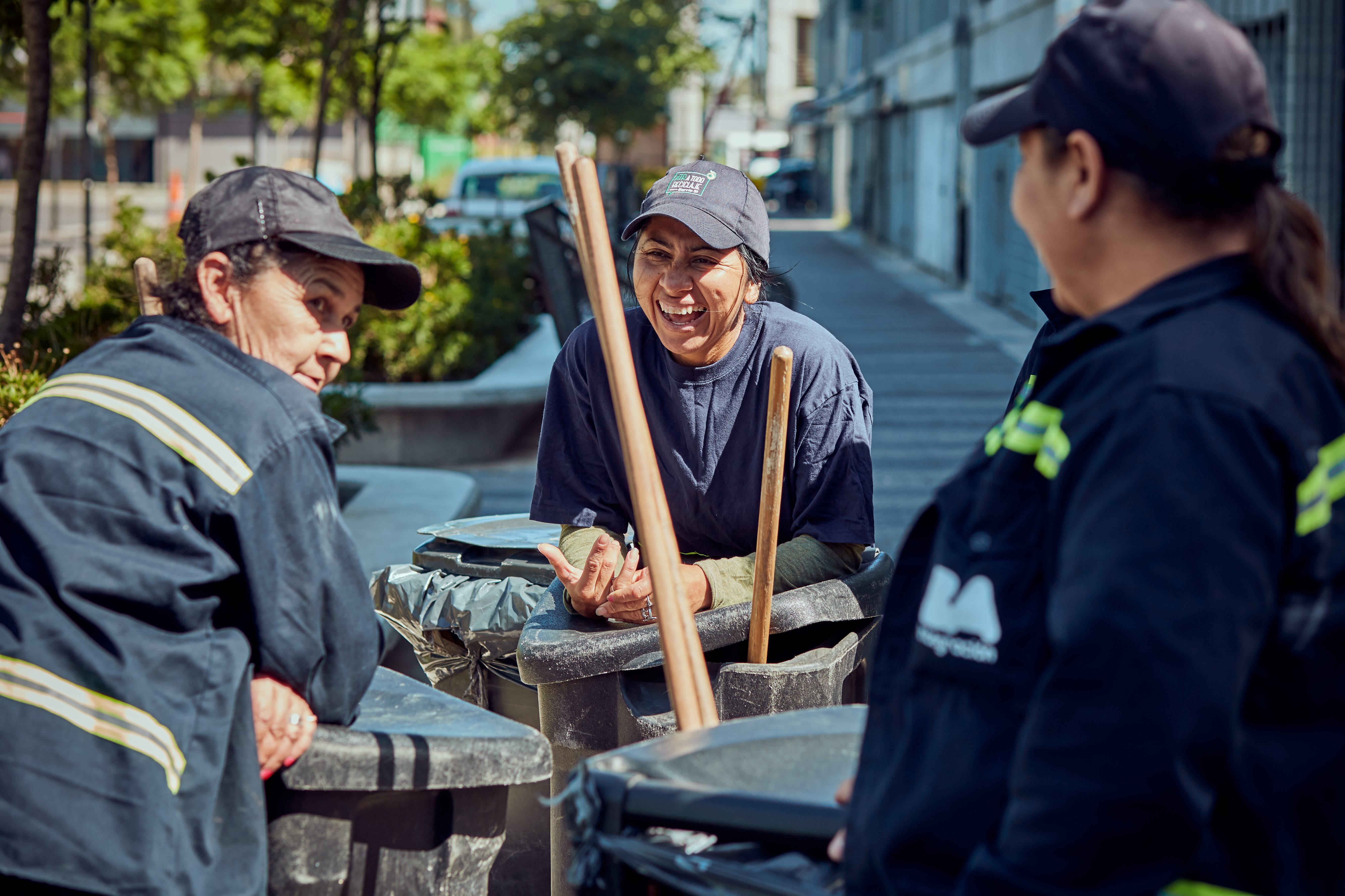 Drei Frauen in grauer Arbeitskleidung inklusive Mütze stehen gut gelaunt im Dreieck zusammen auf einem Bürgersteig vor einer Häuserfront. Jede der Frauen hat eine schwarze, geschlossene, hüfthohe Tonne dabei, welche an der Seite eine Art Halterung haben, aus denen Holzstiele rausschauen. Das Bild deutet darauf hin, dass die Frauen gerade Reinigungsarbeiten auf dieser Straße verrichten.