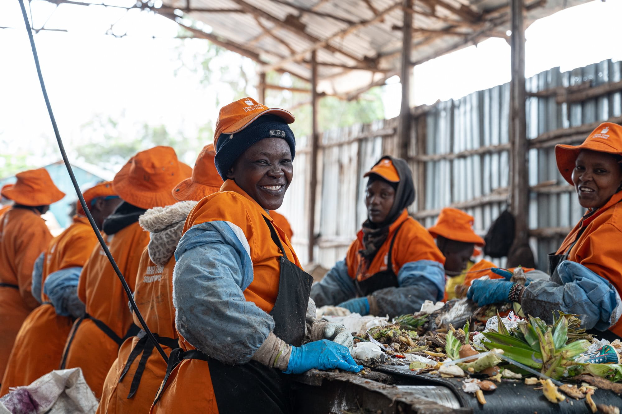 Eine Gruppe von Arbeiterinnen in orangefarbenen Overalls und Mützen, die in einer Recyclinganlage in Kenia Abfälle sortieren. Unter einem Wellblechdach, das von Holzbalken gestützt wird, sind sie damit beschäftigt, verschiedene Arten von Abfallmaterialien auf einem langen Tisch zu trennen. Plastik und organische Materialien sind auf dem Tisch sichtbar.
