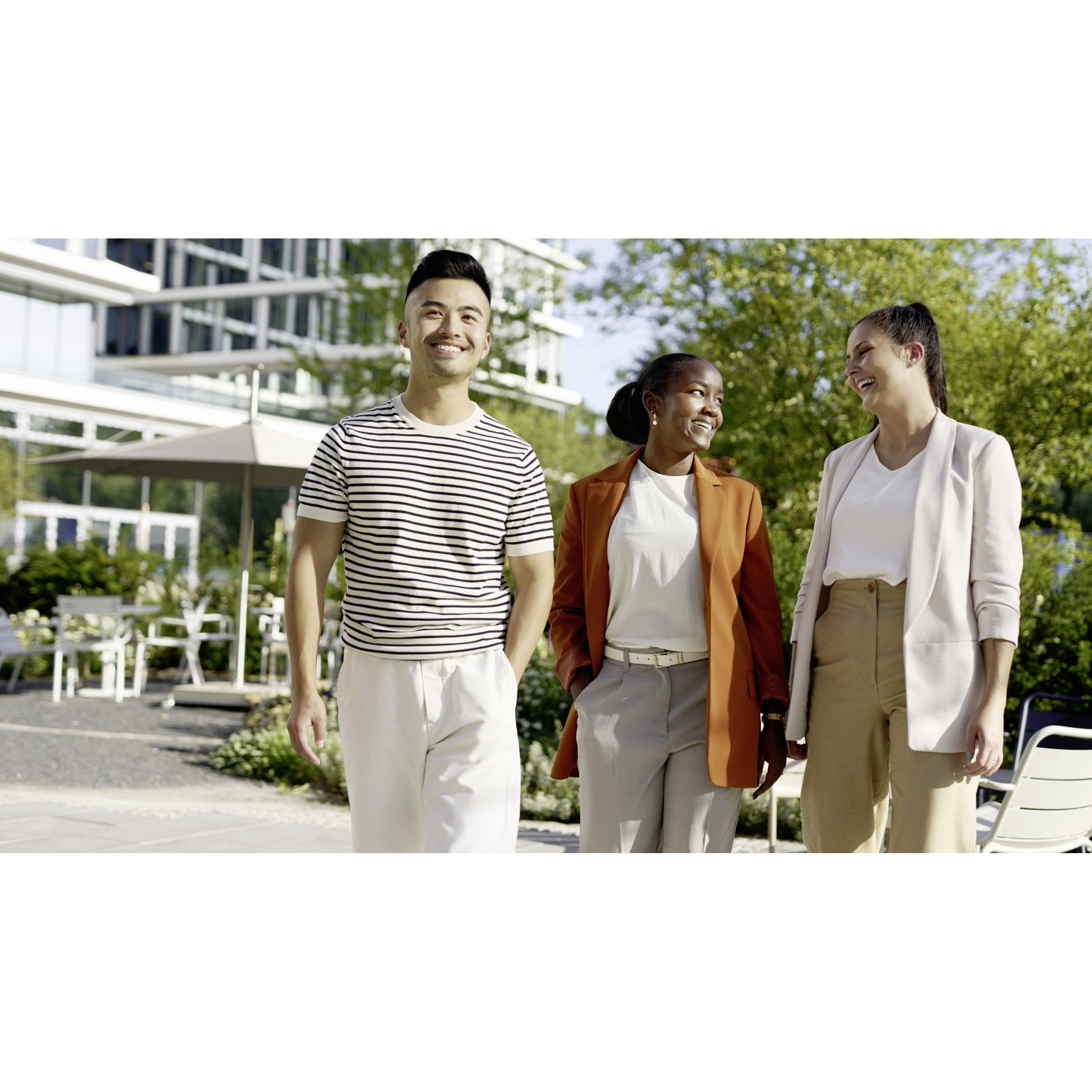 Drei Kolleg*innen gehen an einem sonnigen Tag gemeinsam vor einem Bürogebäude entlang.