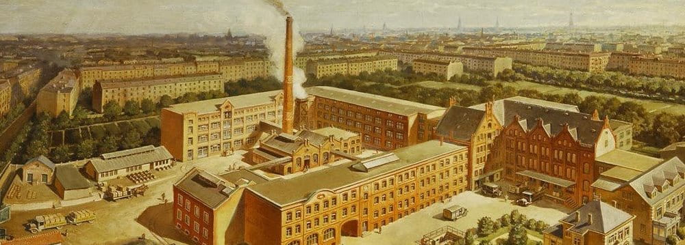 Historic panoramic view of an industrial factory complex with multiple buildings and chimneys, set within an urban landscape.