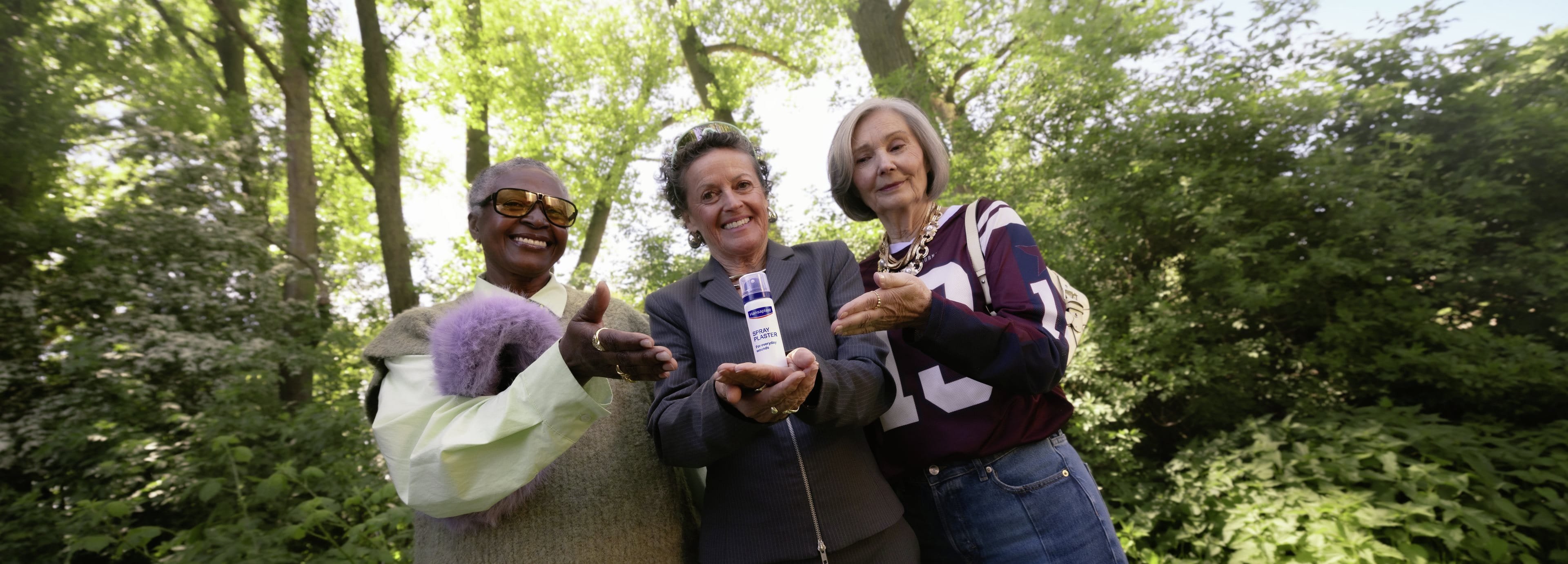 Two older women standing outdoors and smiling, holding and presenting a NIVEA product bottle together, with trees visible in the background.